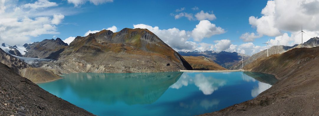 Large size panorama view of Griessee (Gries Lake) and Griesgletscher (Gries Glacier) just across the border between Canton of Ticino and Canton Valais in Switzerland and the international border between Italy and Switzerland in the Lepontine Alps. Near Bedretto Valley and Nufenen mountain pass on the Swiss side and Formazza Valley in Piedmont on the Italian side. Four Wind Turbines visible in the distance on Nufenen mountain pass.