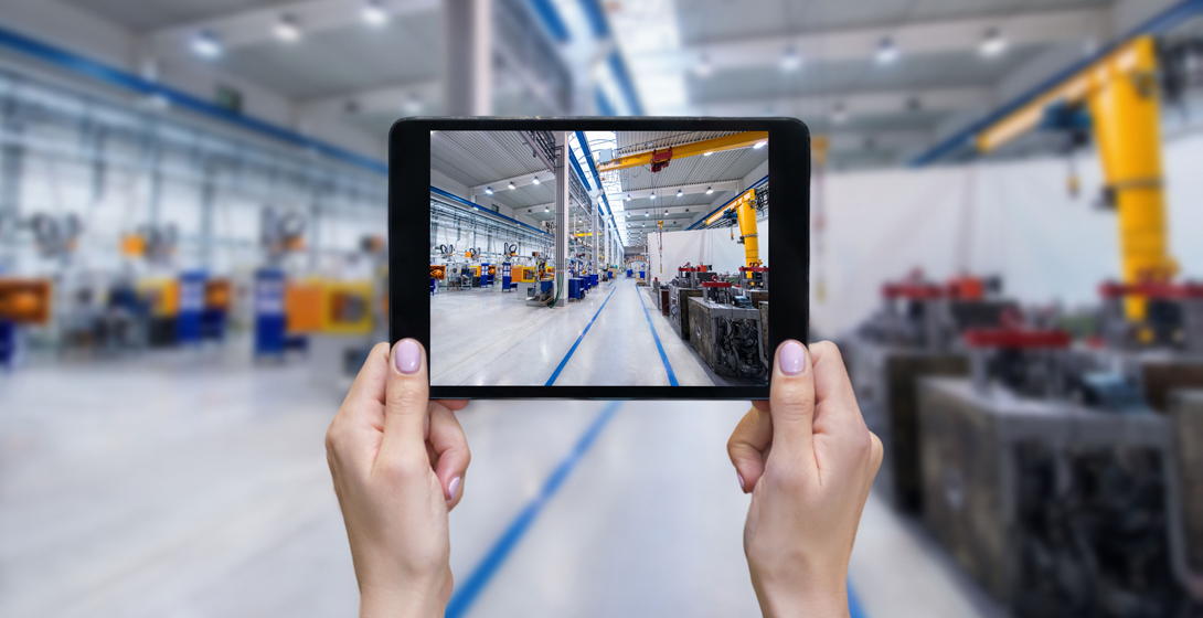Horizontal color image of female hands holding a digital tablet in a modern plastic production line. Ordering on-line from injection moulding factory on a touchscreen tablet computer. Large factory, industrial machines, futuristic robots, crane and manufacturing equipment arranged on clean and shiny flooring in background.